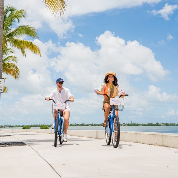 Couple riding complimentary Blue Flamingo bicycles along a palm-lined waterfront path in Key West on a sunny day.