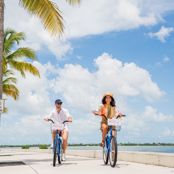 Couple riding complimentary Blue Flamingo bicycles along a palm-lined waterfront path in Key West on a sunny day.