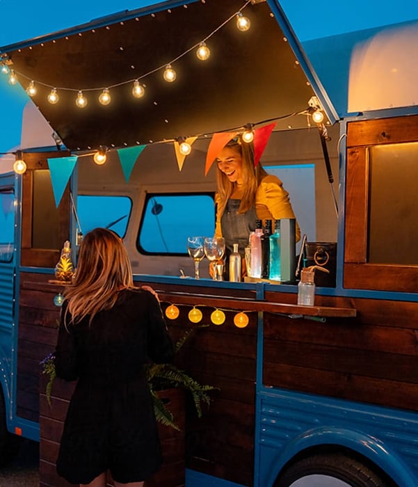A woman serves a patron at a food truck illuminated with string lights, at dusk
