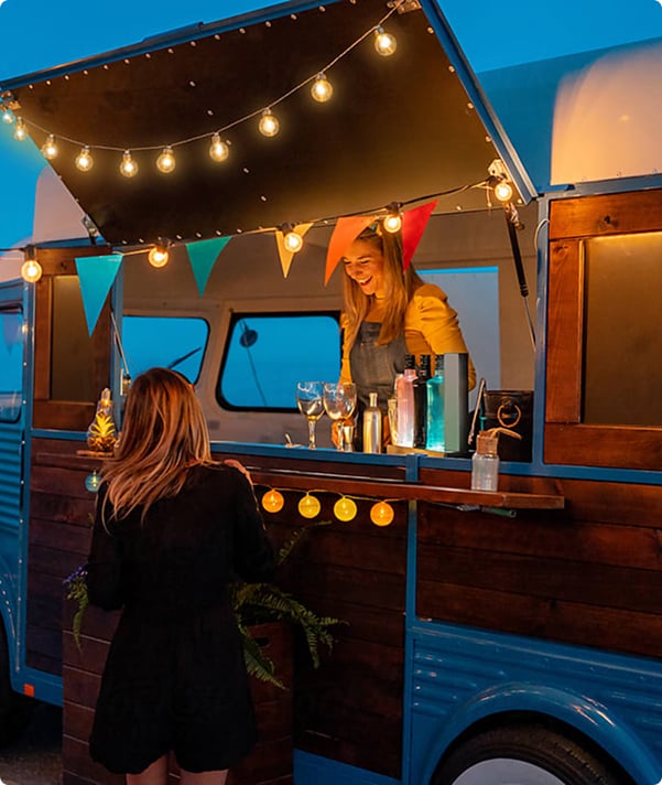 A woman serves a patron at a food truck illuminated with string lights, at dusk