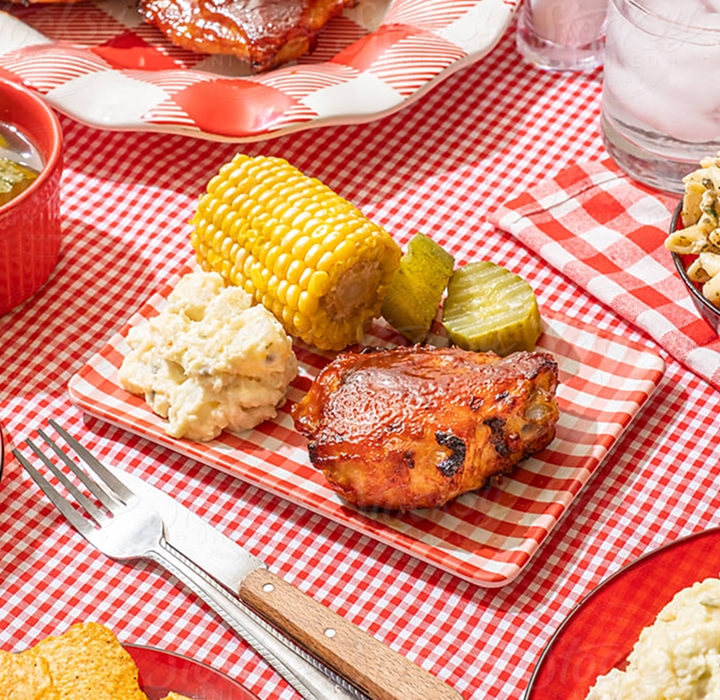 A platter with grilled corn, potato salad and chicken on a red checkboard cloth