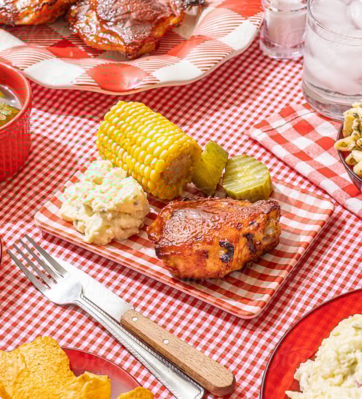 A platter with grilled corn, potato salad and chicken on a red checkboard cloth