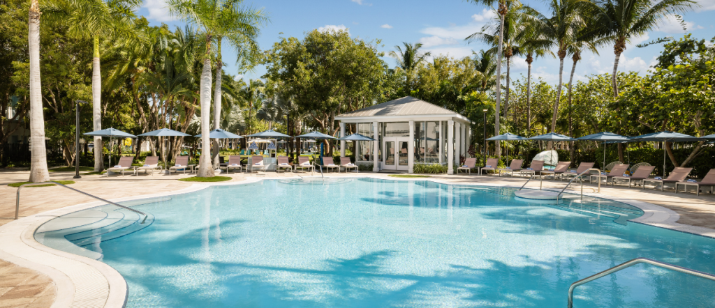 resort pool at Blue Flamingo Key West surrounded by palm trees, lounge chairs, blue umbrellas, and a glass-walled fitness center