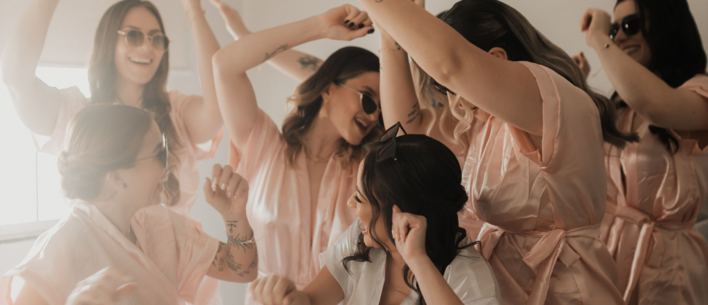 Four friends celebrating a bachelorette party in a decorated hotel room with pink balloons and party props